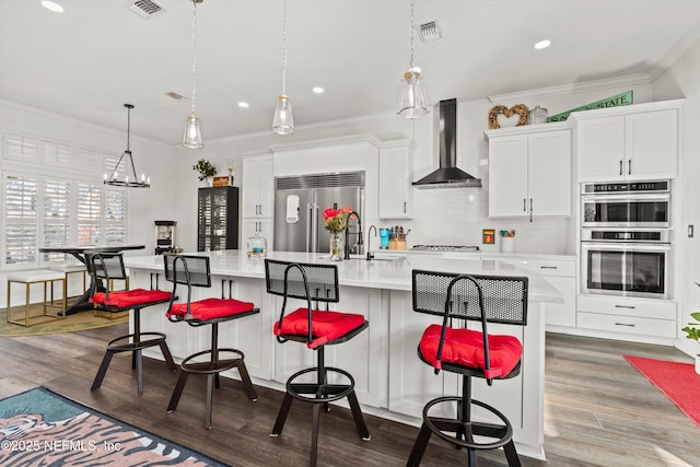 kitchen featuring white cabinets, a spacious island, wall chimney range hood, appliances with stainless steel finishes, and a kitchen bar