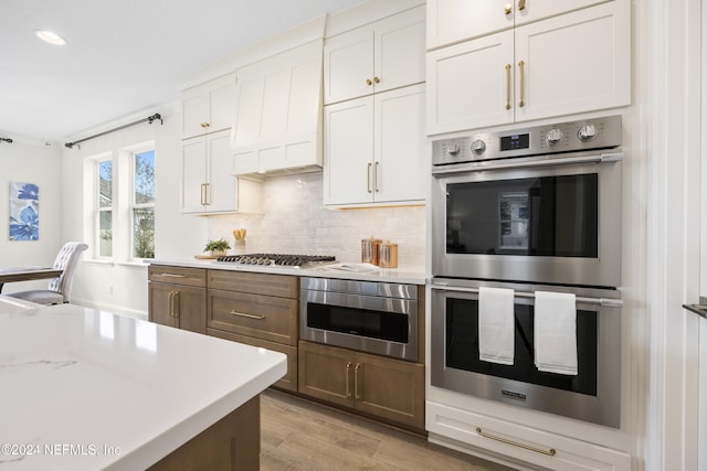 kitchen with light stone countertops, custom range hood, stainless steel appliances, light hardwood / wood-style flooring, and white cabinetry