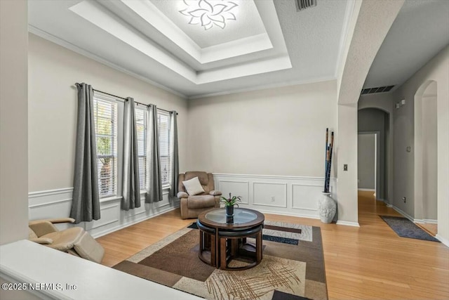 sitting room with a raised ceiling, light wood-type flooring, a decorative wall, visible vents, and arched walkways