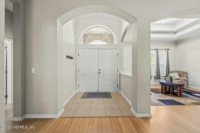entrance foyer with a tray ceiling, ornamental molding, baseboards, arched walkways, and light wood finished floors