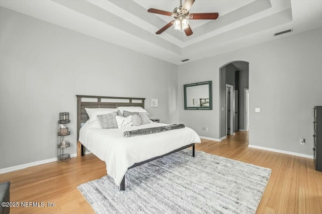 bedroom featuring a tray ceiling, light wood finished floors, baseboards, visible vents, and arched walkways