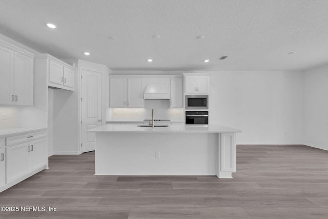 kitchen with a center island with sink, tasteful backsplash, stainless steel microwave, wall oven, and white cabinets