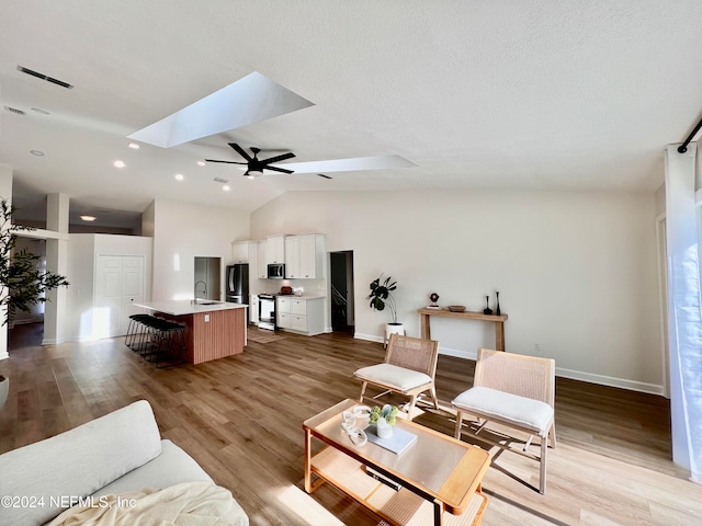 living room featuring sink, light hardwood / wood-style flooring, ceiling fan, and vaulted ceiling with skylight