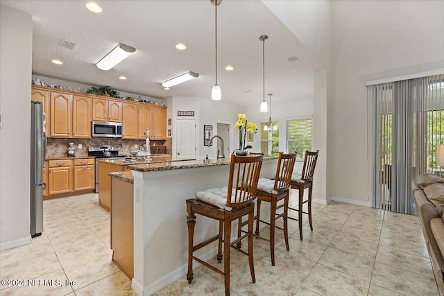 kitchen featuring a breakfast bar area, hanging light fixtures, appliances with stainless steel finishes, dark stone countertops, and a center island with sink