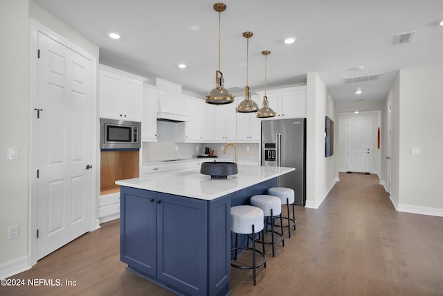 kitchen featuring dark wood-type flooring, hanging light fixtures, an island with sink, appliances with stainless steel finishes, and white cabinetry