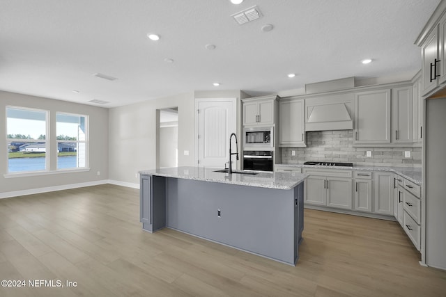 kitchen with light stone countertops, light hardwood / wood-style floors, a center island with sink, a water view, and black appliances