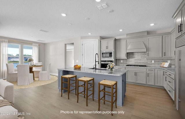 kitchen with gray cabinetry, a center island with sink, appliances with stainless steel finishes, light hardwood / wood-style floors, and light stone counters