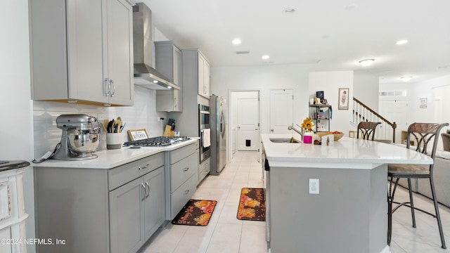 kitchen featuring wall chimney exhaust hood, gray cabinetry, a breakfast bar, stainless steel gas stovetop, and an island with sink