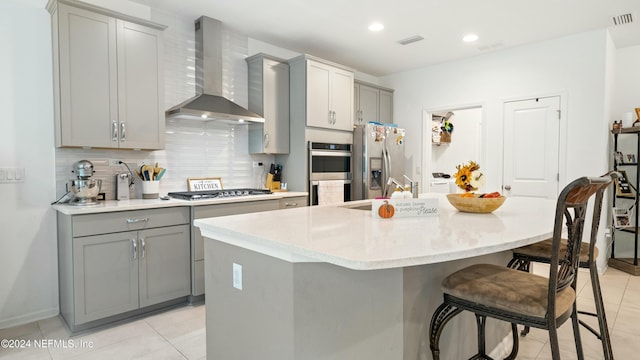 kitchen featuring an island with sink, stainless steel appliances, gray cabinetry, and wall chimney range hood