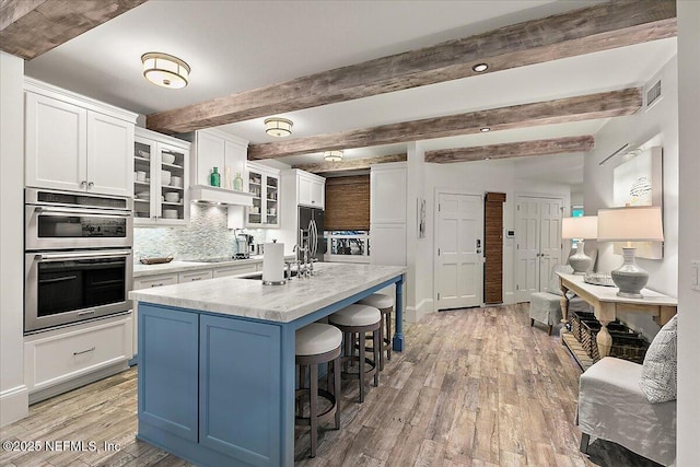 kitchen featuring an island with sink, backsplash, beamed ceiling, and white cabinetry