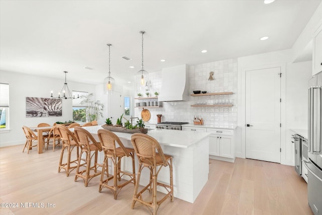 kitchen with custom exhaust hood, light hardwood / wood-style flooring, white cabinets, hanging light fixtures, and an island with sink