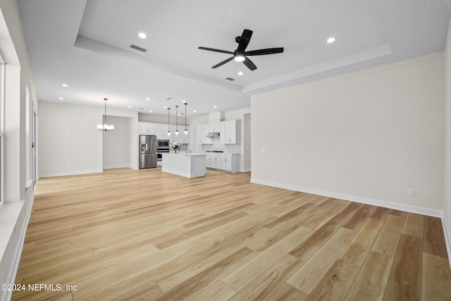 unfurnished living room featuring ceiling fan with notable chandelier, light wood-type flooring, and a tray ceiling