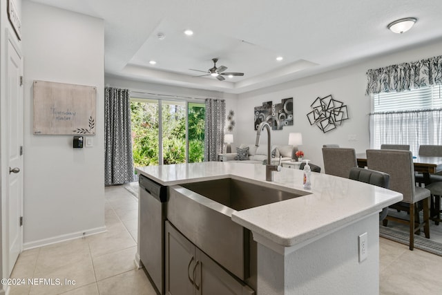 kitchen featuring dishwasher, a center island with sink, sink, ceiling fan, and a tray ceiling