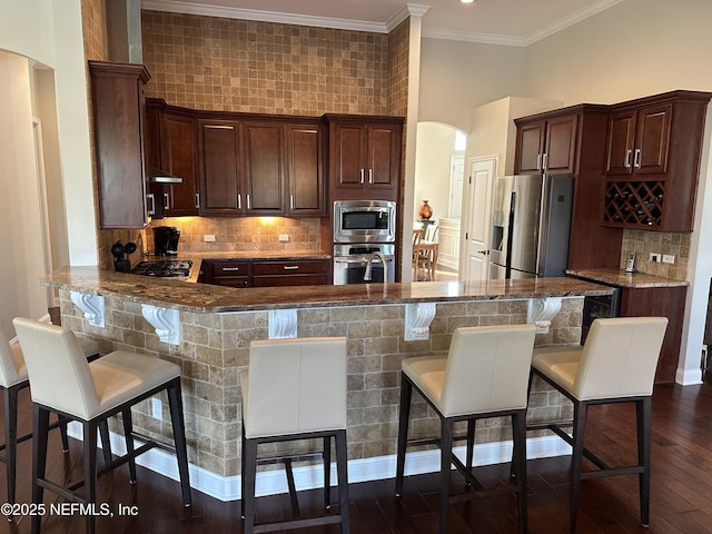 kitchen featuring arched walkways, a breakfast bar area, appliances with stainless steel finishes, light stone countertops, and a peninsula