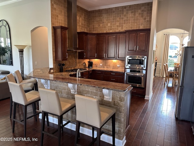 kitchen with arched walkways, appliances with stainless steel finishes, a peninsula, wall chimney range hood, and a kitchen bar