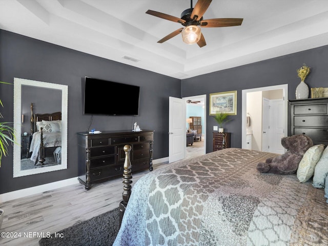 bedroom with a tray ceiling, connected bathroom, ceiling fan, and light wood-type flooring