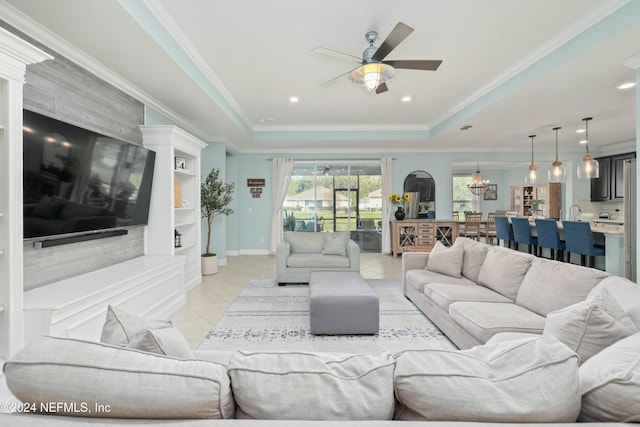 living room with ceiling fan with notable chandelier, a tray ceiling, crown molding, light tile patterned floors, and built in features