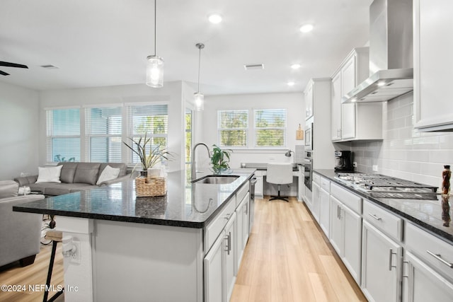 kitchen with a center island with sink, white cabinets, wall chimney exhaust hood, appliances with stainless steel finishes, and light hardwood / wood-style floors