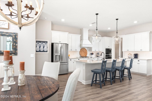 kitchen featuring stainless steel refrigerator with ice dispenser, premium range hood, decorative light fixtures, a center island with sink, and white cabinets