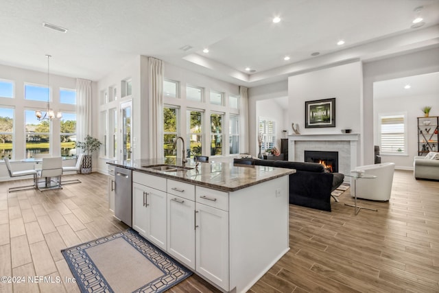 kitchen with white cabinetry, stainless steel dishwasher, dark stone counters, a tiled fireplace, and a kitchen island with sink