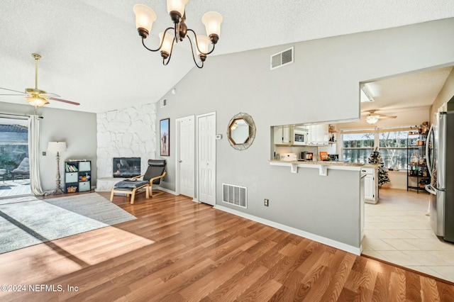 living room with light hardwood / wood-style flooring, high vaulted ceiling, a textured ceiling, a fireplace, and ceiling fan with notable chandelier