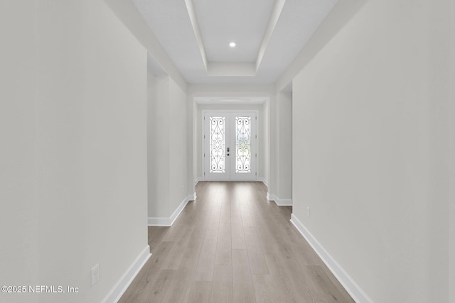 corridor featuring french doors, light wood-style flooring, baseboards, a tray ceiling, and recessed lighting