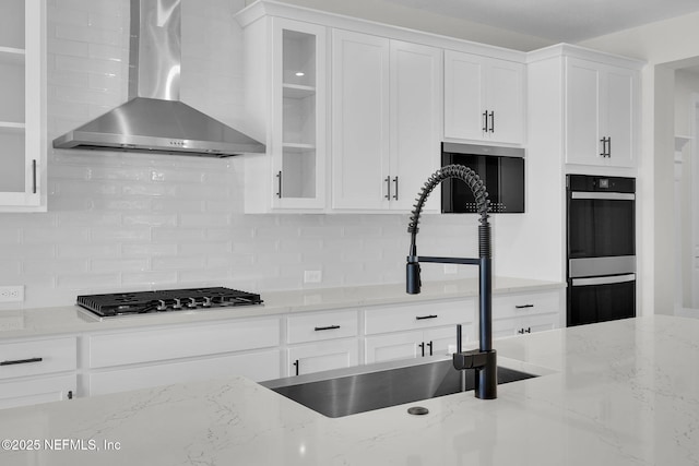 kitchen with wall chimney exhaust hood, stainless steel gas stovetop, tasteful backsplash, white cabinets, and glass insert cabinets