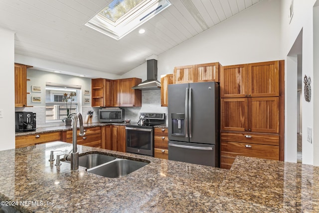 kitchen featuring appliances with stainless steel finishes, vaulted ceiling with skylight, dark stone countertops, and wall chimney range hood