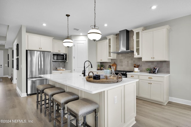 kitchen featuring decorative backsplash, appliances with stainless steel finishes, wall chimney exhaust hood, a center island with sink, and light hardwood / wood-style floors