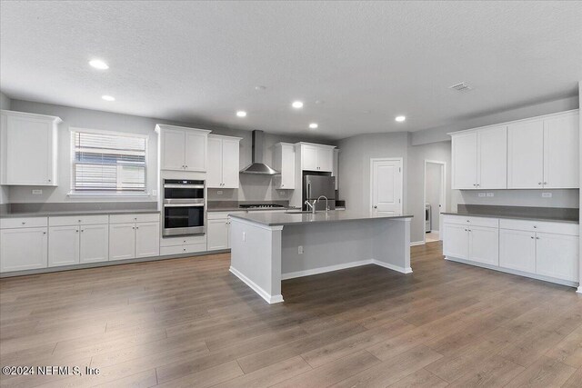 kitchen with a kitchen island with sink, white cabinets, light hardwood / wood-style flooring, wall chimney exhaust hood, and appliances with stainless steel finishes