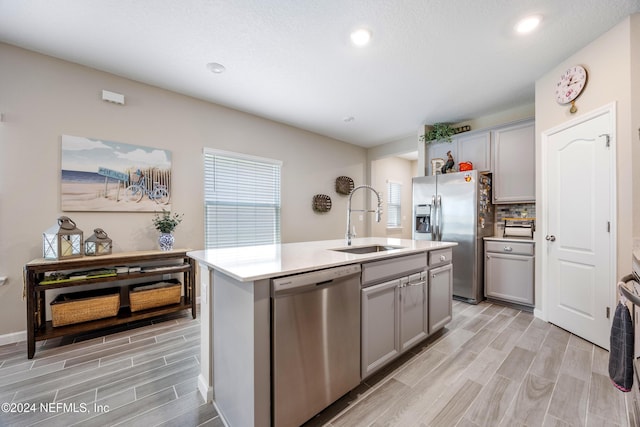 kitchen with appliances with stainless steel finishes, light wood-type flooring, a kitchen island with sink, sink, and gray cabinets