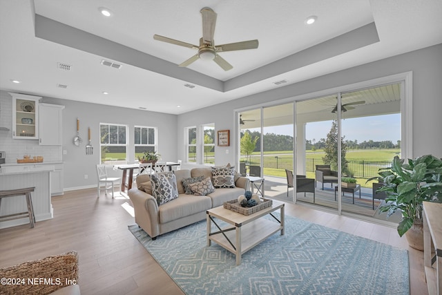 living room with a raised ceiling, ceiling fan, and light wood-type flooring