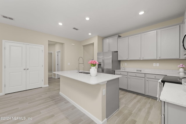 kitchen featuring stainless steel fridge, sink, light stone countertops, gray cabinets, and a center island with sink