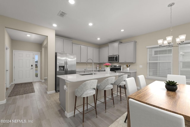 kitchen with appliances with stainless steel finishes, sink, a kitchen island with sink, a notable chandelier, and light wood-type flooring