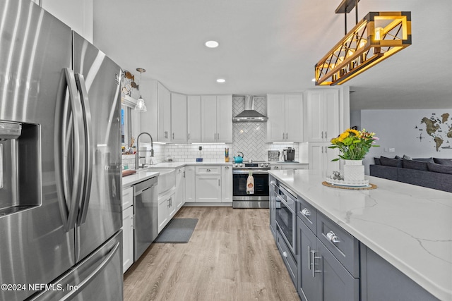 kitchen with light hardwood / wood-style flooring, wall chimney exhaust hood, appliances with stainless steel finishes, decorative light fixtures, and white cabinetry