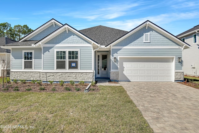 craftsman-style house featuring a shingled roof, stone siding, an attached garage, decorative driveway, and a front lawn