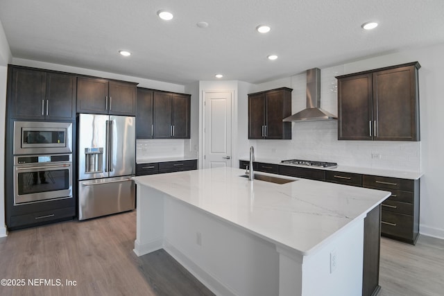 kitchen featuring appliances with stainless steel finishes, light wood-style floors, a kitchen island with sink, a sink, and wall chimney range hood