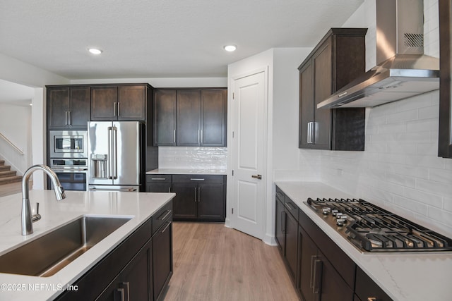 kitchen with stainless steel appliances, a sink, wall chimney range hood, dark brown cabinetry, and light stone countertops