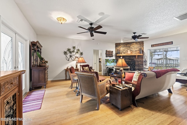 living room with ceiling fan, french doors, a stone fireplace, a textured ceiling, and light wood-type flooring