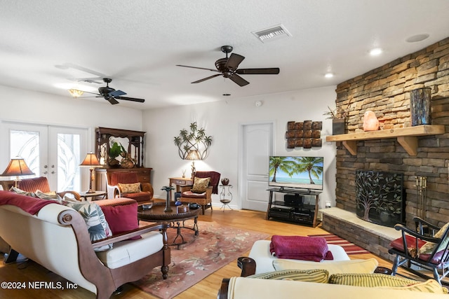 living room featuring french doors, a stone fireplace, light hardwood / wood-style flooring, ceiling fan, and a textured ceiling