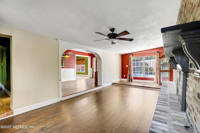 unfurnished living room with wood finished floors, a fireplace with raised hearth, arched walkways, ceiling fan, and a textured ceiling