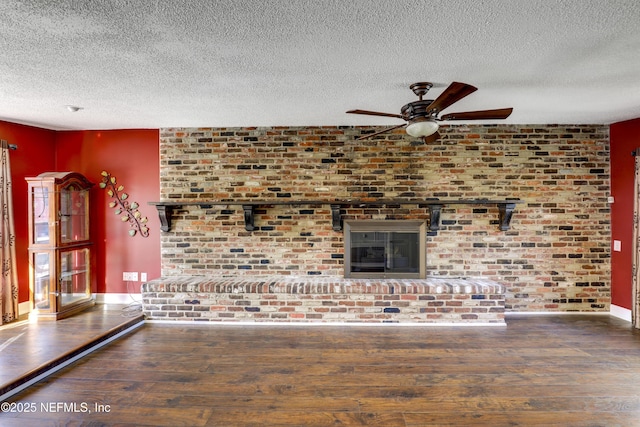 unfurnished living room featuring a textured ceiling, wood finished floors, baseboards, a brick fireplace, and ceiling fan