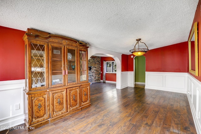 unfurnished dining area featuring arched walkways, a wainscoted wall, a textured ceiling, and wood finished floors