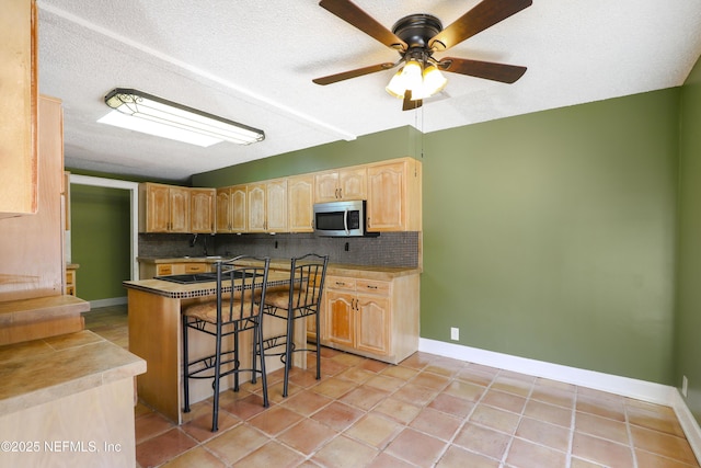 kitchen featuring a breakfast bar area, light brown cabinets, a textured ceiling, stainless steel microwave, and tasteful backsplash