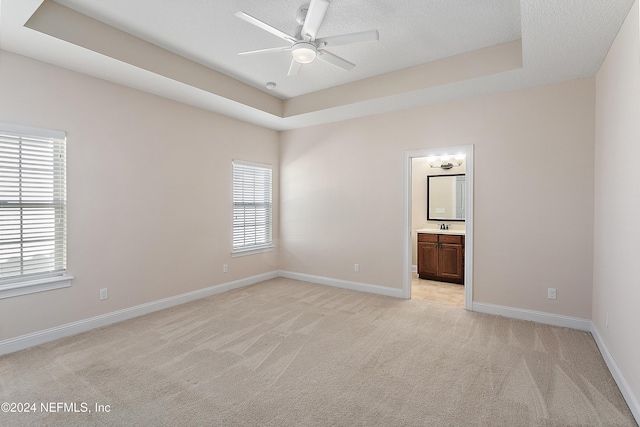 unfurnished bedroom featuring ensuite bathroom, a textured ceiling, light colored carpet, a raised ceiling, and ceiling fan