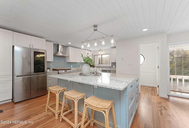 kitchen with wall chimney range hood, hanging light fixtures, appliances with stainless steel finishes, light hardwood / wood-style floors, and white cabinetry