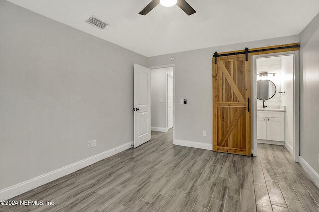 unfurnished bedroom with ensuite bathroom, a barn door, ceiling fan, and light hardwood / wood-style flooring