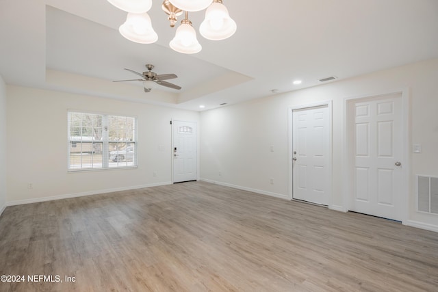 empty room with a tray ceiling, light hardwood / wood-style flooring, and ceiling fan with notable chandelier