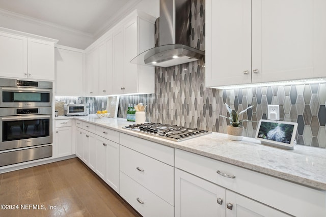 kitchen with appliances with stainless steel finishes, ornamental molding, wall chimney range hood, wood-type flooring, and white cabinets