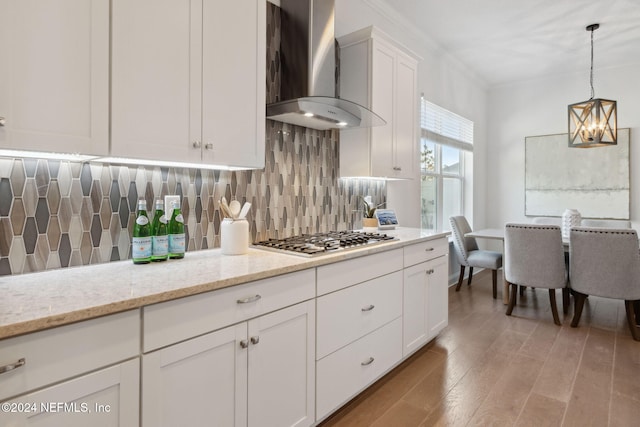 kitchen with light stone countertops, stainless steel gas cooktop, wall chimney range hood, decorative light fixtures, and white cabinetry